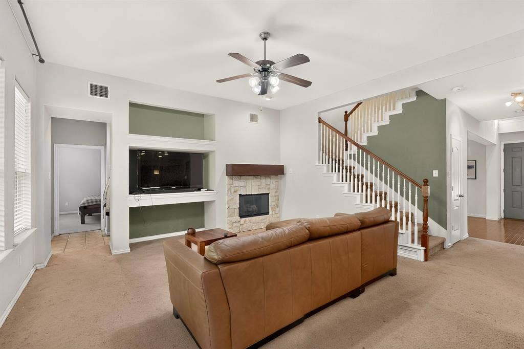 Living area featuring a floor-to-ceiling stone-faced fireplace with a wooden mantel, integrated wall shelving, a central ceiling fan, and a staircase with a wooden handrail