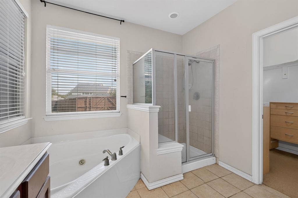 Bathroom featuring a corner soaking tub with deck-mounted faucet, a separate glass-enclosed shower, neutral tile flooring, and windows with blinds