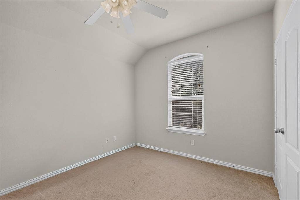 Neutral-toned room featuring a vaulted ceiling, arched-top window with integrated blinds, wall-mounted ceiling fan, and carpeted flooring