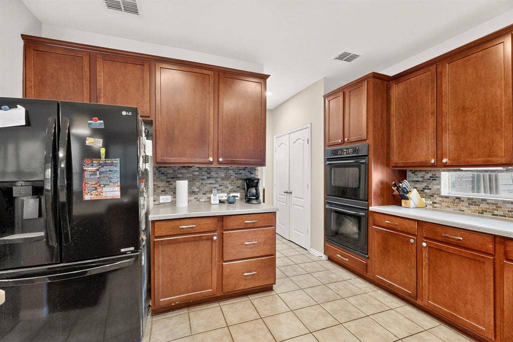 Kitchen featuring stained wood cabinetry, stone-tiled backsplash, built-in double wall oven, black refrigerator, and tile flooring