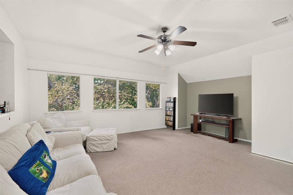 Spacious room featuring neutral carpeting, a multi-pane ceiling fan with integrated lighting, and a trio of large windows with exterior foliage
