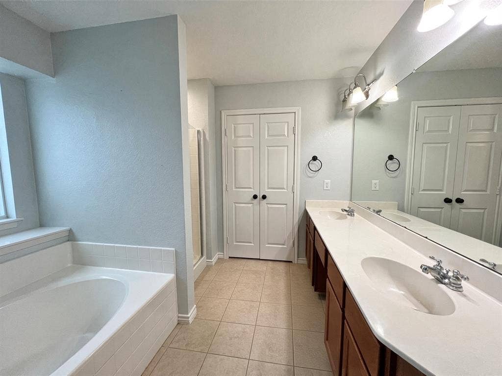 Dual vanity with a stone-surface countertop, porcelain sinks, tile flooring, soaking tub with tile surround, and expansive wall-mounted mirror