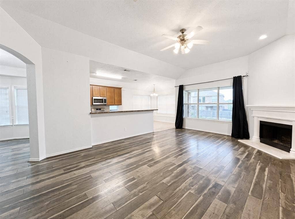 Living area featuring wood-finish flooring, vaulted ceilings, a fireplace with a white mantel, a ceiling fan, and a breakfast bar