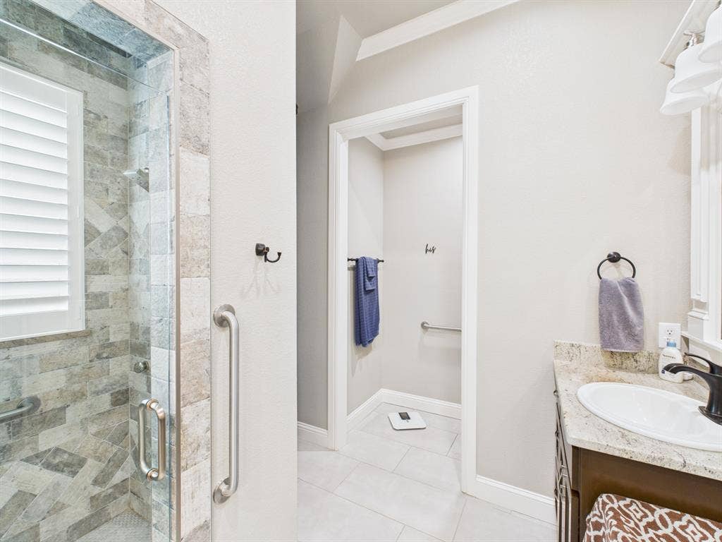 1201 E Iberian Court, Granbury, TX 76048 Walk-in shower featuring multi-tonal stone tile surround and glass door, stone-surface vanity with undermount sink, dark-finish cabinetry, and matte black fixtures