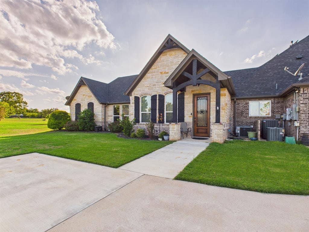 1201 E Iberian Court, Granbury, TX 76048 Stone and brick facade featuring a gabled roofline, decorative timber framing over the entrance, wood-finish front door with glass insert, and dark shutters