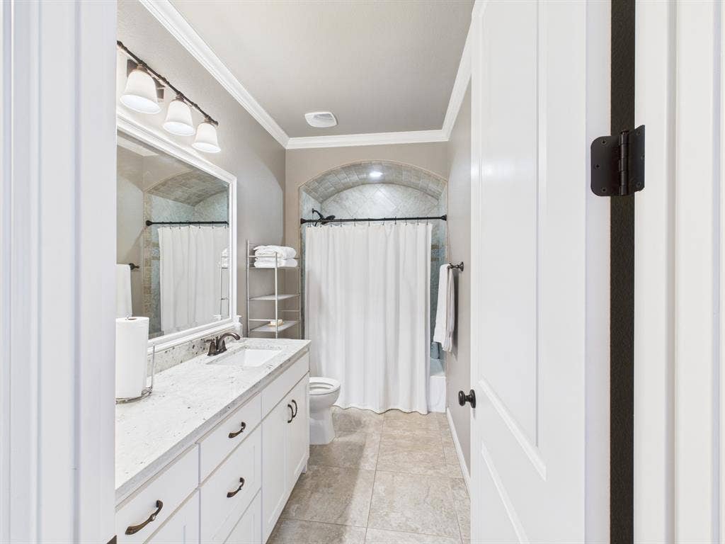 1201 E Iberian Court, Granbury, TX 76048 Bathroom featuring a stone-topped vanity with undermount sink, multi-drawer white cabinetry, crown molding, tile flooring, and a tiled arched shower alcove