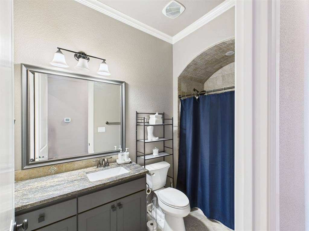 1201 E Iberian Court, Granbury, TX 76048 Bathroom featuring a charcoal vanity with a stone countertop, an integrated rectangular sink, a framed vanity mirror with a three-light fixture, and a tiled shower alcove with an arched ceiling detail