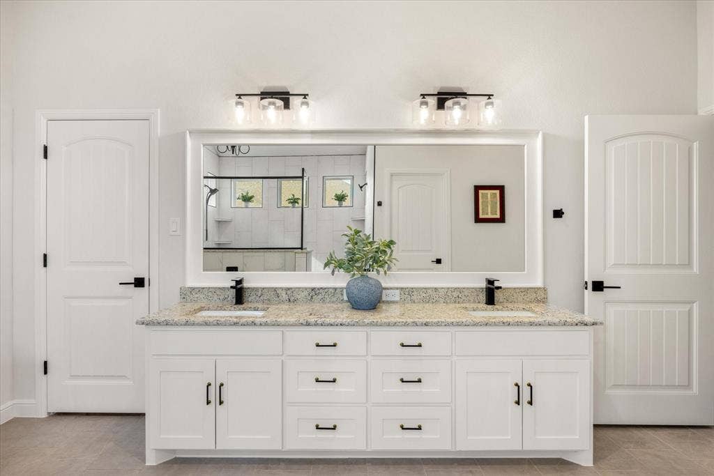 Dual-sink vanity featuring stone countertop, white cabinetry with matte black hardware, large framed mirror, and dual wall-mounted light fixtures