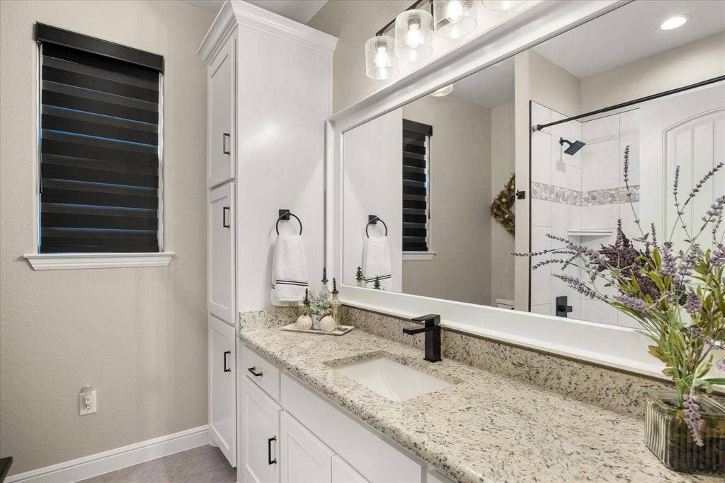 Bathroom featuring white cabinetry with a floor-to-ceiling storage tower, stone countertop, matte black faucet, full-width framed mirror, and walk-in shower with tiled surround
