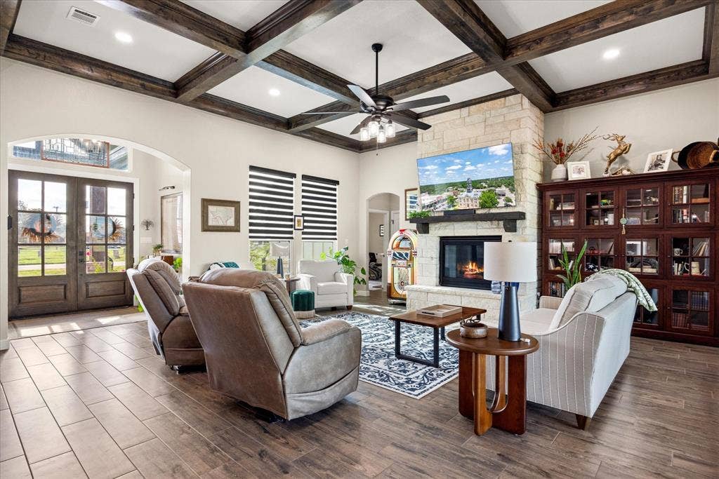 Living area featuring a coffered ceiling with wood-finish beams, a stone-faced fireplace with a timber mantel, tile flooring, and built-in glass-front cabinetry
