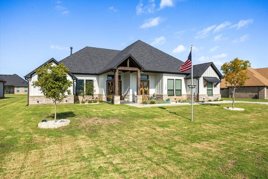 Modern farmhouse exterior featuring vertical white siding, stone veneer base, dark metal roof accents, and a covered entryway with timber-frame details