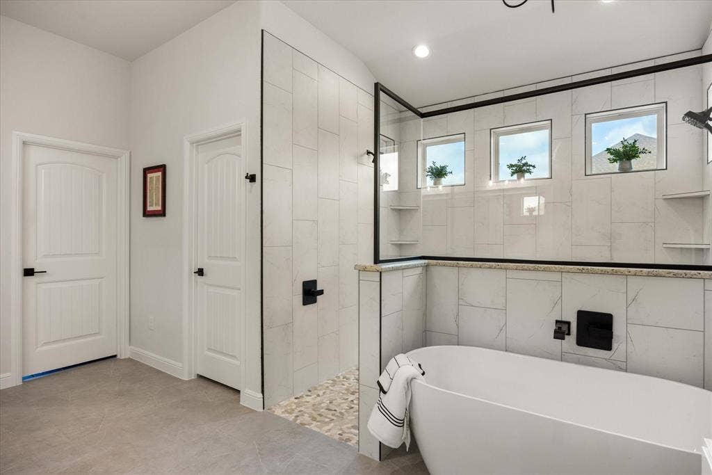 Freestanding soaking tub with matte black wall-mounted faucet, walk-in shower featuring a pebble-stone floor, glass enclosure with black framing, and neutral tile wall surrounds
