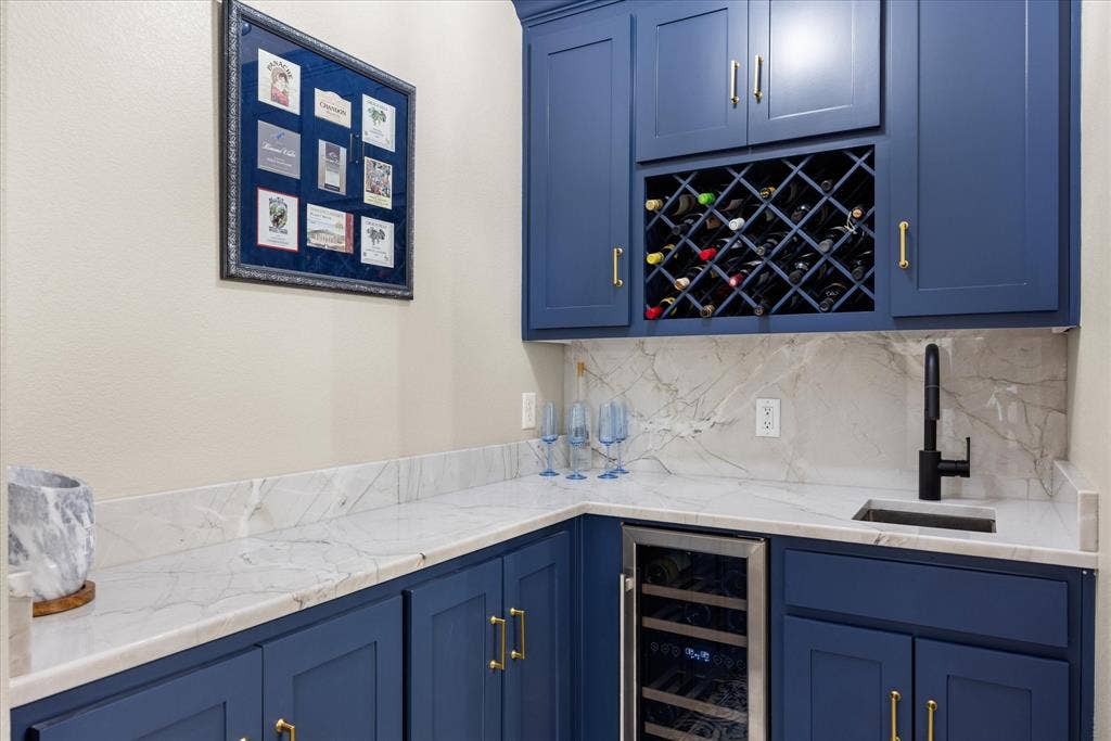 Custom blue cabinetry featuring built-in wine storage, stone-finish countertops, full-height stone-finish backsplash, and an integrated beverage refrigerator with a matte black faucet