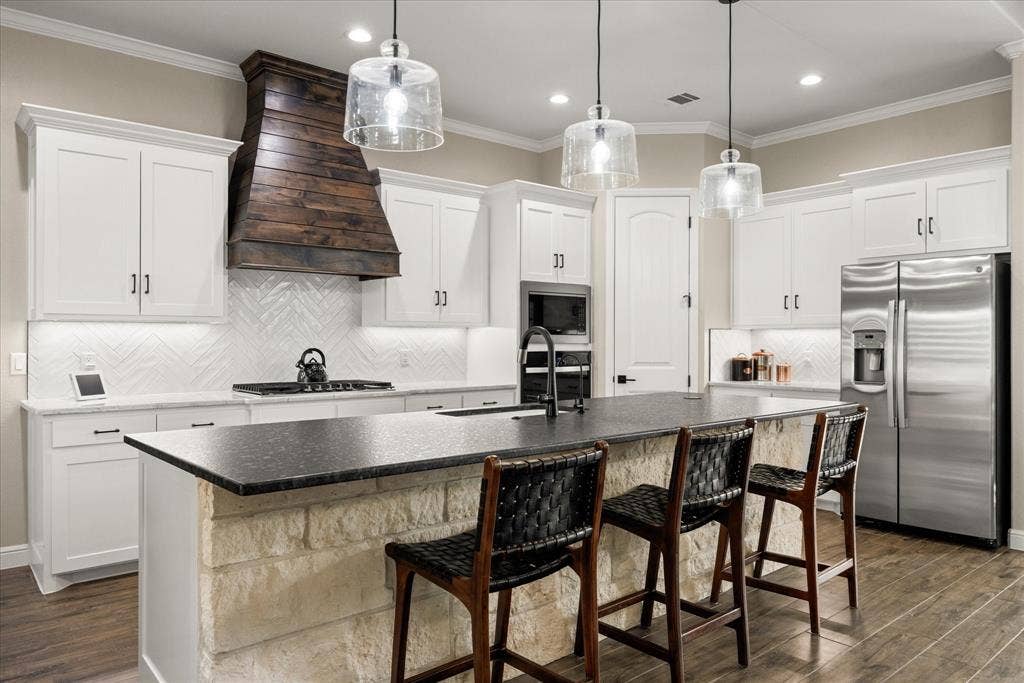 Kitchen featuring white cabinetry, a natural stone-faced island with a dark countertop, a custom dark wood range hood, a herringbone-patterned tile backsplash, and stainless steel appliances