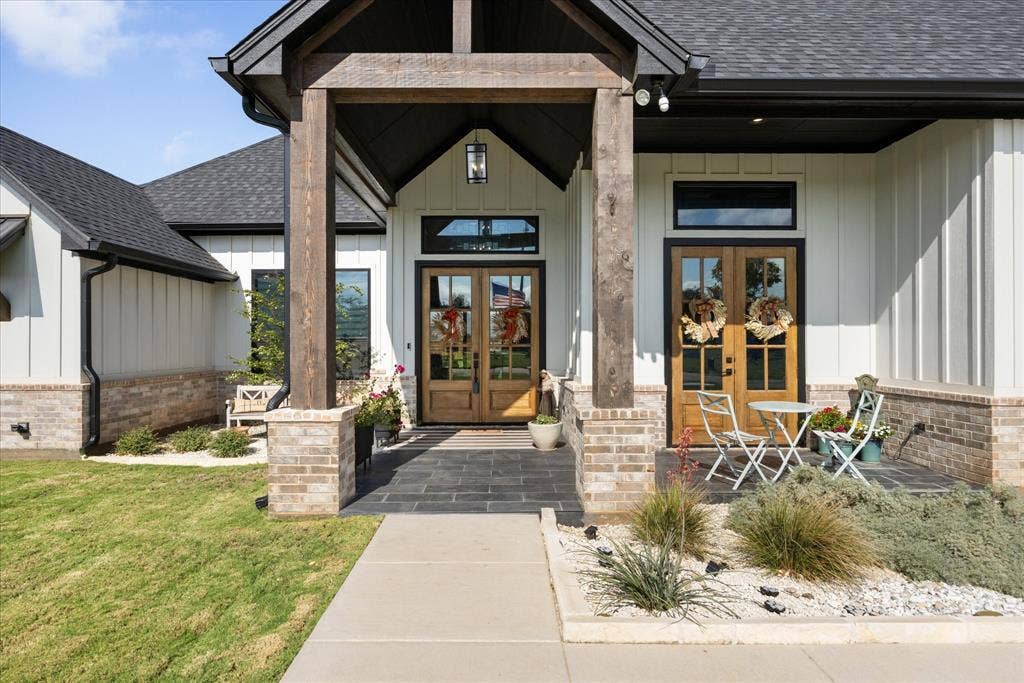 Board-and-batten siding exterior featuring dual wood-frame glass entry doors, stone veneer accents, timber-frame porch supports, and a dark stone-tile patio