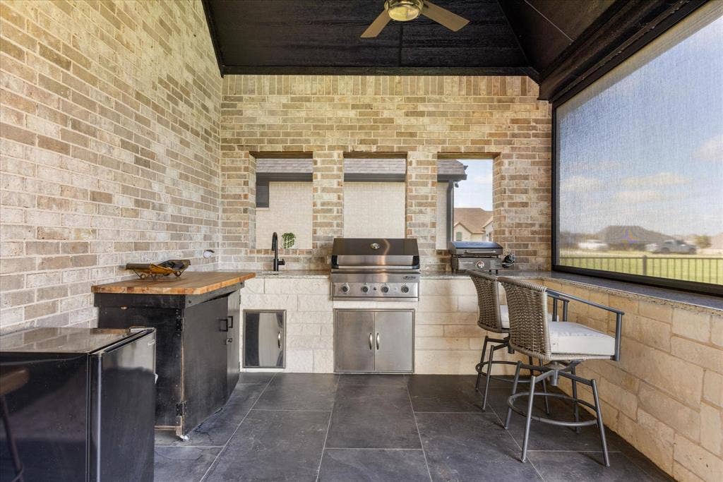 Covered outdoor kitchen featuring brick masonry walls, stone-surfaced countertops, a built-in stainless steel grill, a mounted ceiling fan, and large-format dark tile flooring