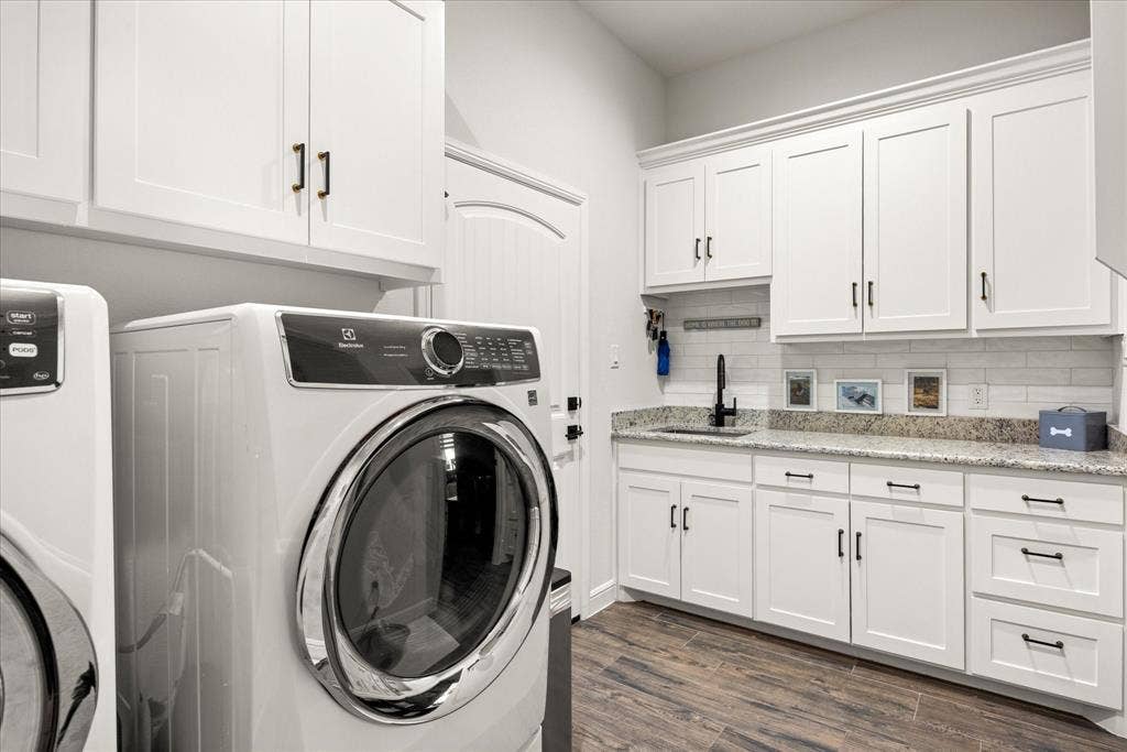 Laundry room featuring white Shaker-style cabinetry, stone-finish countertops, undermount sink with matte black faucet, white subway tile backsplash, and wood-finish flooring