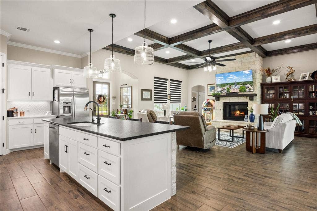 Kitchen and living area featuring coffered ceilings with exposed beams, wood-finish flooring, a kitchen island with stone base, and a stone-clad fireplace with a built-in mantle