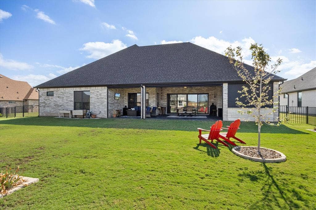 Rear exterior featuring a stone masonry facade, covered patio, dark shingle roof, and a fenced backyard with a landscaped tree island