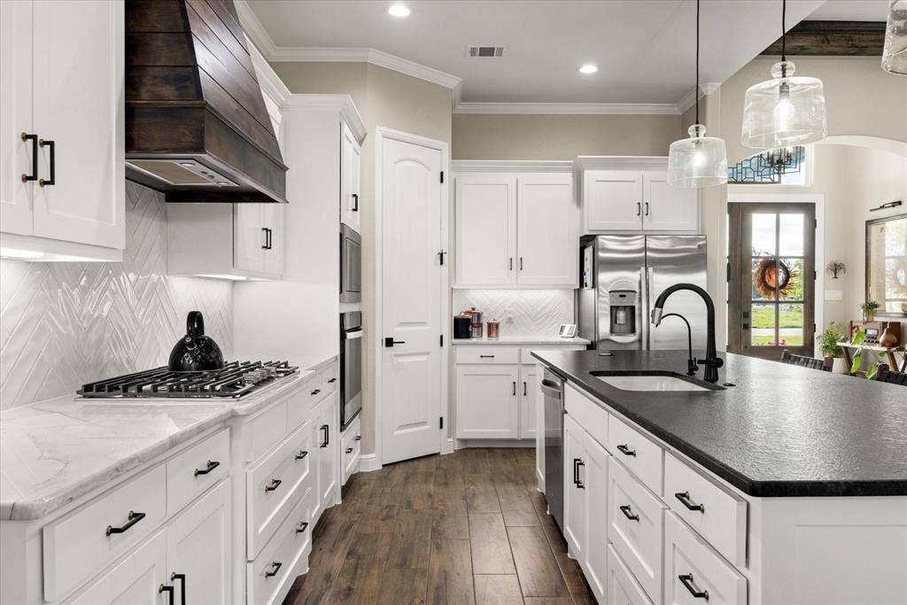 Kitchen featuring custom white cabinetry, a dark-toned wood-finish hood range, a large center island with a dark countertop, white herringbone-patterned backsplash, and wood-finish flooring