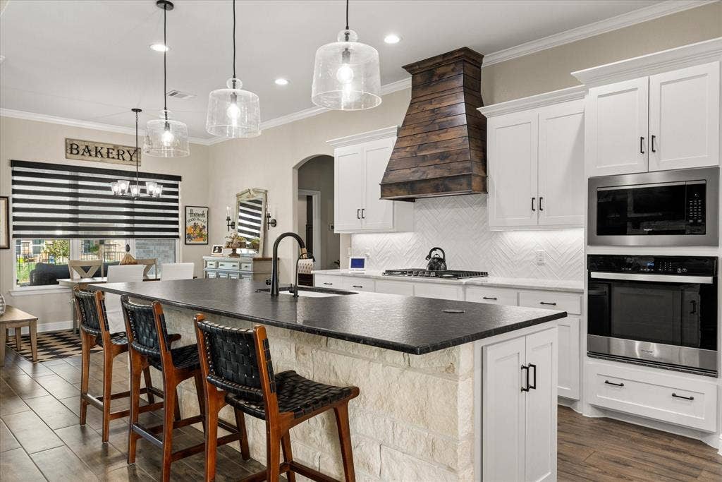 Kitchen featuring an expansive center island with a stone base and dark countertop, white cabinetry, herringbone-patterned backsplash, wood-finish vent hood, and built-in stainless steel double ovens