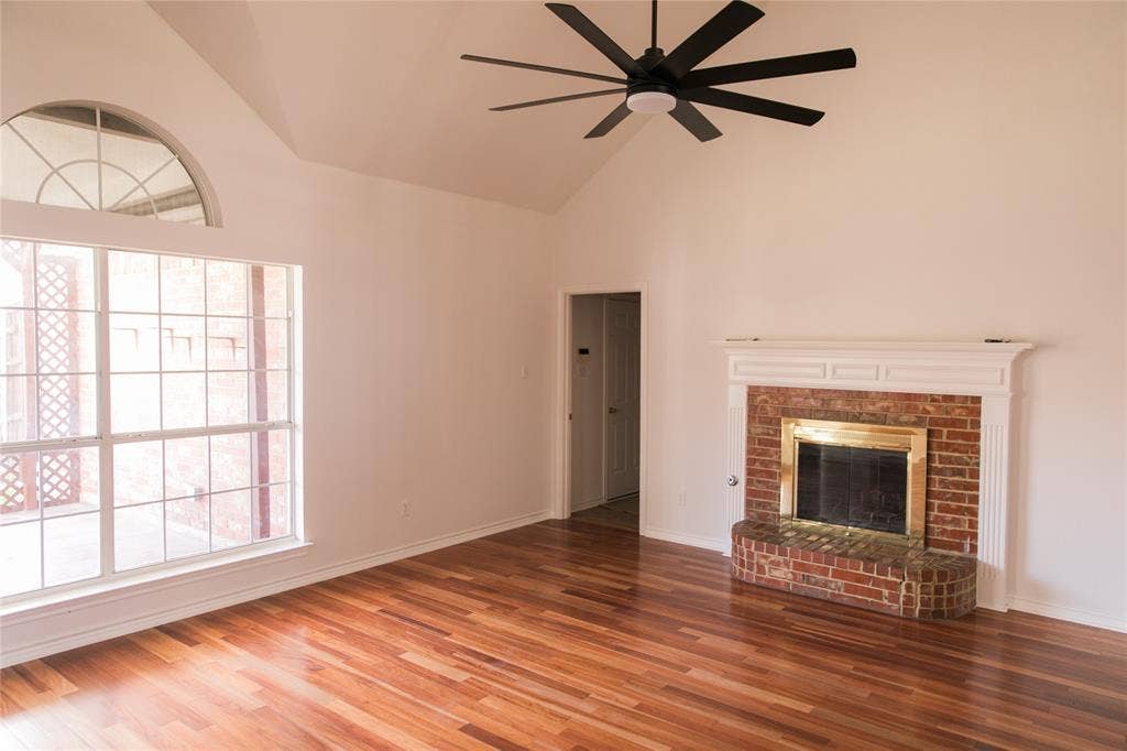 404 Belmont Court, Lewisville, TX 75067 Vaulted ceiling with a matte black multi-blade ceiling fan, wood-finish flooring, brick fireplace with a white mantel, and an expansive window featuring an arched transom
