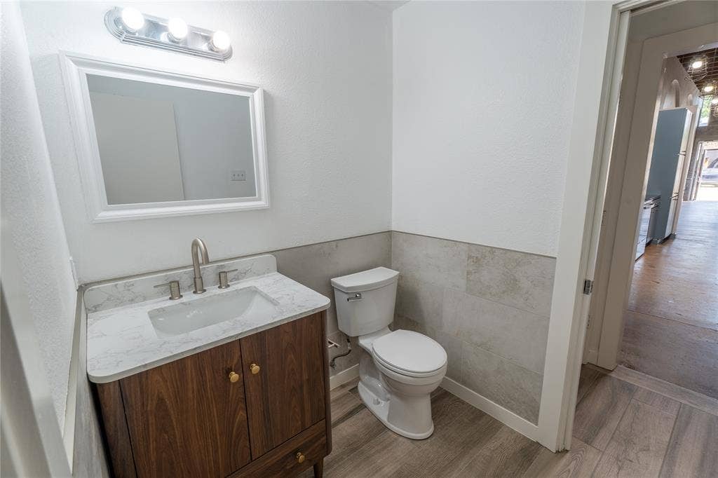 3012 Canton Street, Dallas, TX 75226 Vanity featuring a stone-patterned countertop, under-mount sink, and wood-finish cabinetry, complemented by wall-mounted lighting, a rectangular mirror, and neutral tile wainscoting