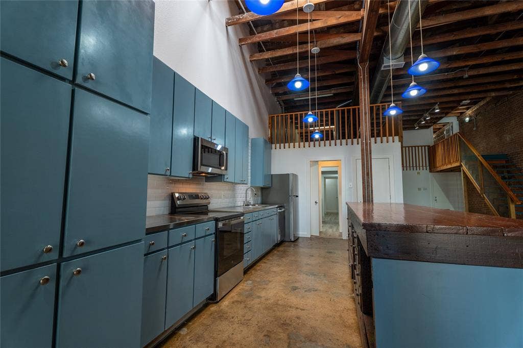 3012 Canton Street, Dallas, TX 75226 Industrial-style kitchen featuring exposed wood ceiling joists, stained concrete flooring, blue cabinetry, stainless steel appliances, and a built-in island with a dark wood countertop