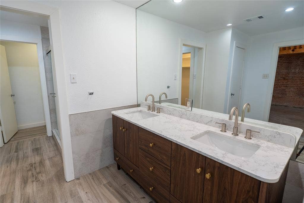 3012 Canton Street, Dallas, TX 75226 Double-sink vanity featuring a stone-composite countertop, wood-finish cabinetry, metallic fixtures, and a wall-to-wall mirror, complemented by wood-finish flooring and large-format wall tile