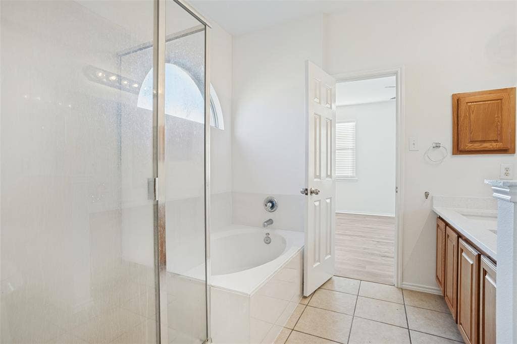 1507 Bankston Drive, Wylie, TX 75098 Bathroom featuring a glass-enclosed shower, a deep soaking tub, an arched transom window, a built-in wooden vanity with countertop, and ceramic tile flooring
