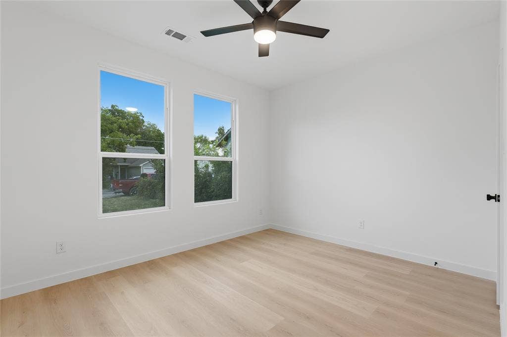 Bright room featuring wood-finish flooring, white walls, dual windows, and a matte black ceiling fan with integrated lighting