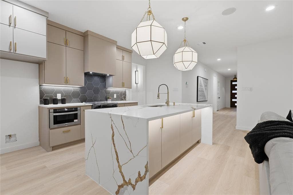 Kitchen featuring a waterfall-edge island with stone-finish countertop, dual geometric pendant lighting, custom cabinetry with integrated under-cabinet lighting, hexagonal tile backsplash, and wood-finish flooring