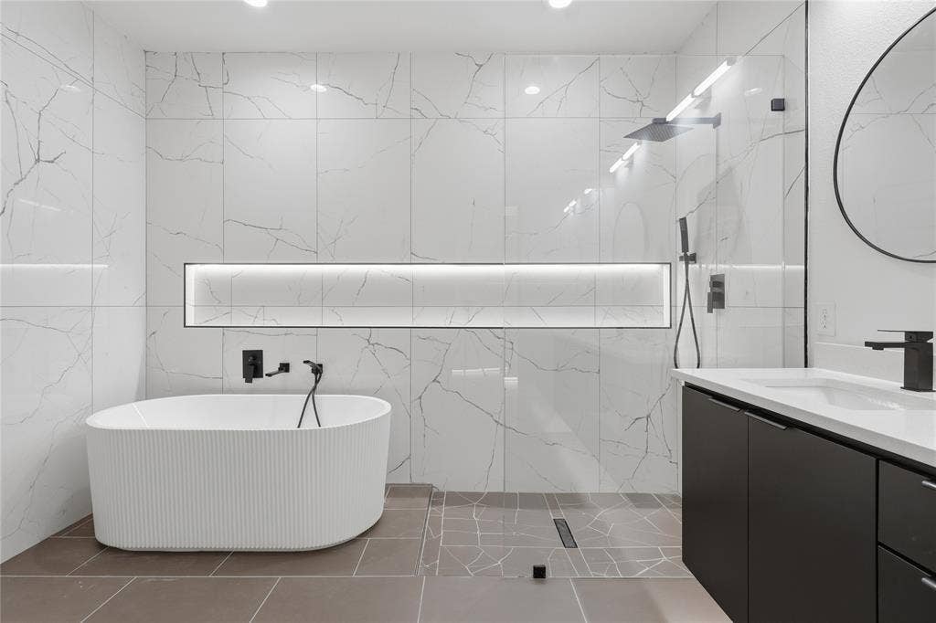 Freestanding fluted soaking tub with wall-mounted matte black fixtures, walk-in shower enclosed by a glass panel, recessed linear lighting niche, and a custom vanity with a contrasting cabinet finish