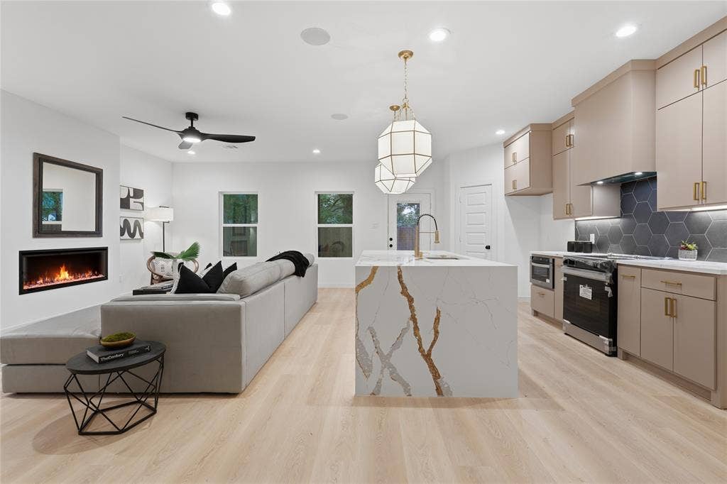 Kitchen and living area featuring wood-finish flooring, a central waterfall-edge island with stone-finish countertops, geometric pendant lighting, a recessed linear fireplace, and cabinetry with matte gold hardware