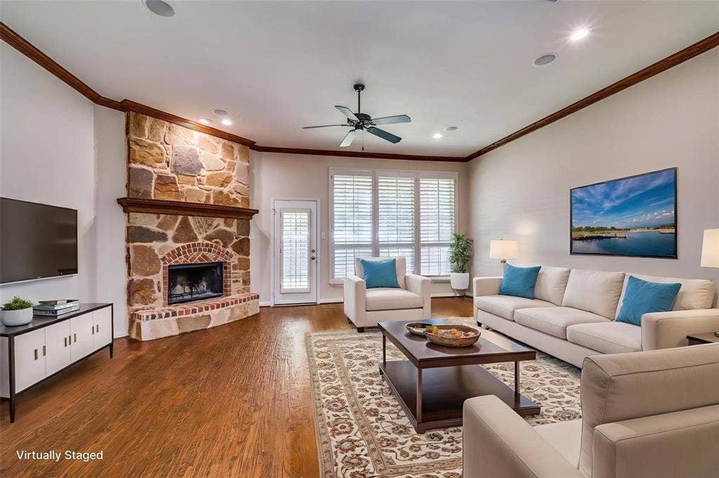 1441 Kirkwood Lane, Prosper, TX 75078 Living area featuring wood-finish flooring, full-height stone fireplace with a brick surround, crown molding, plantation shutters, and a ceiling fan