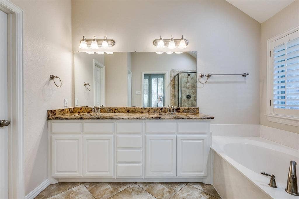 1441 Kirkwood Lane, Prosper, TX 75078 Dual vanity featuring white cabinetry, stone-surface countertop, dual under-mount sinks, wall-to-wall mirror with dual three-light fixtures, and a soaking tub with tile flooring
