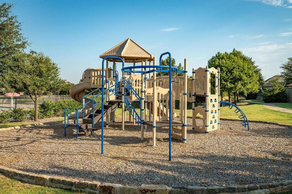 1441 Kirkwood Lane, Prosper, TX 75078 Recreational playground featuring a multi-level structure with slides, climbing walls, overhead bars, and a covered canopy, situated on a stone-bordered gravel ground cover
