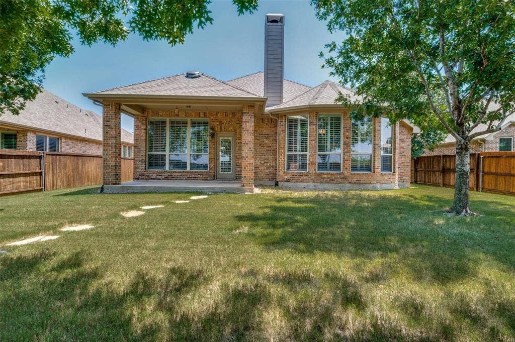 1441 Kirkwood Lane, Prosper, TX 75078 Brick exterior featuring a covered patio, tall chimney, expansive windows with grid detailing, and a fenced backyard with stone pavers