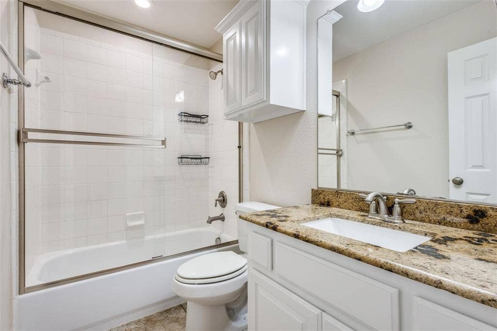 1441 Kirkwood Lane, Prosper, TX 75078 Bathroom featuring a granite-topped vanity with an integrated basin, white shaker-style cabinetry, a wall-mounted storage cabinet, and a tiled tub-shower combination with a sliding glass enclosure