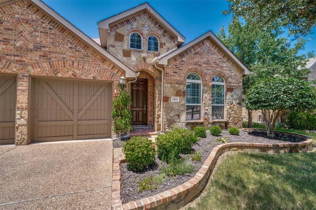 1441 Kirkwood Lane, Prosper, TX 75078 Brick and stone charming exterior featuring arched window detailing, carriage-style garage doors, and a curved masonry garden border