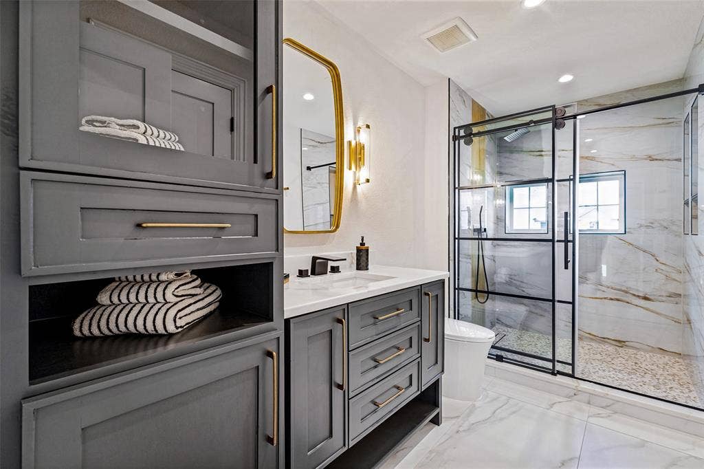 4604 Devon Drive, Parker, TX 75002 Custom gray cabinetry with brass-finish hardware, quartz countertop, arched wall-mounted mirror, and a walk-in shower featuring a sliding black-frame glass door and stone-style flooring