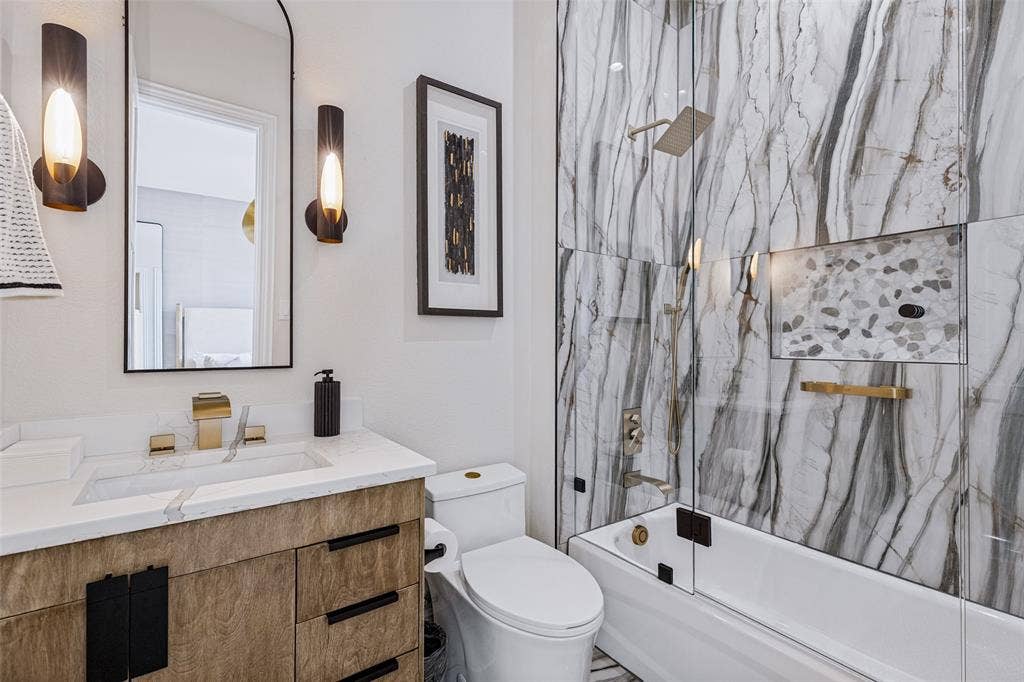 4604 Devon Drive, Parker, TX 75002 Bathroom featuring a wood-finish vanity with a rectangular sink, wall-mounted gold-tone faucet, arched mirror, and floor-to-ceiling veined tile shower surround with a built-in recessed niche