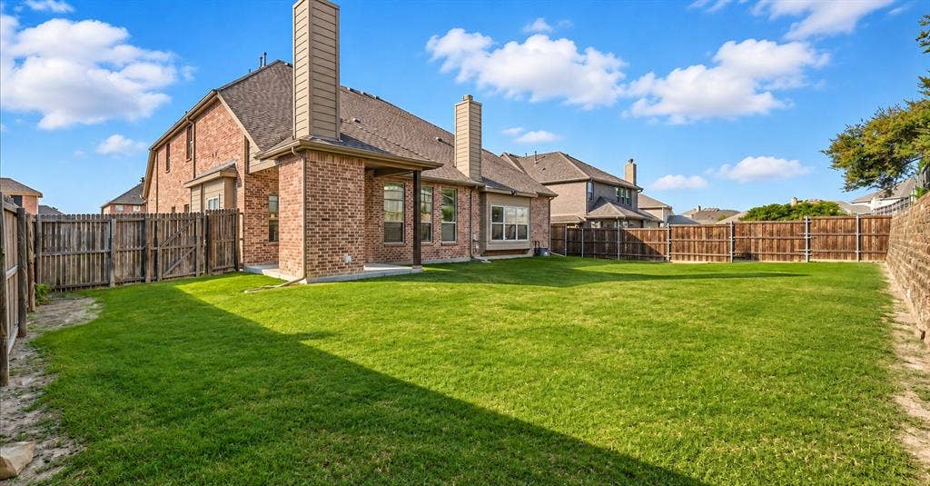 632 Grisham Drive, Fate, TX 75087 Backyard featuring a brick exterior, two masonry chimneys, a covered patio, and perimeter wood fencing surrounding a grass lawn