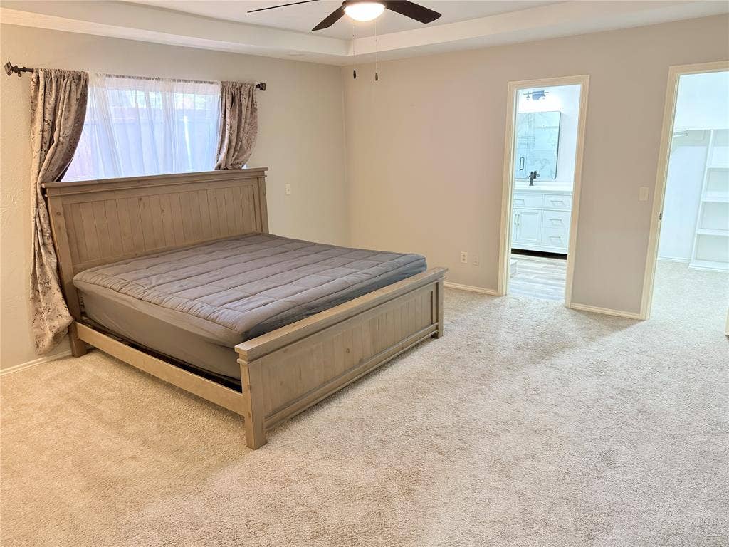 Bedroom featuring a tray ceiling, wall-to-wall carpeting, and a ceiling fan, with entryways revealing an en-suite bathroom vanity and a built-in shelving unit