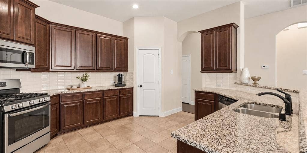 632 Grisham Drive, Fate, TX 75087 Kitchen featuring dark wood cabinetry, stone countertops, stainless steel appliances, a tile backsplash, and a double-basin undermount sink with a pull-down faucet