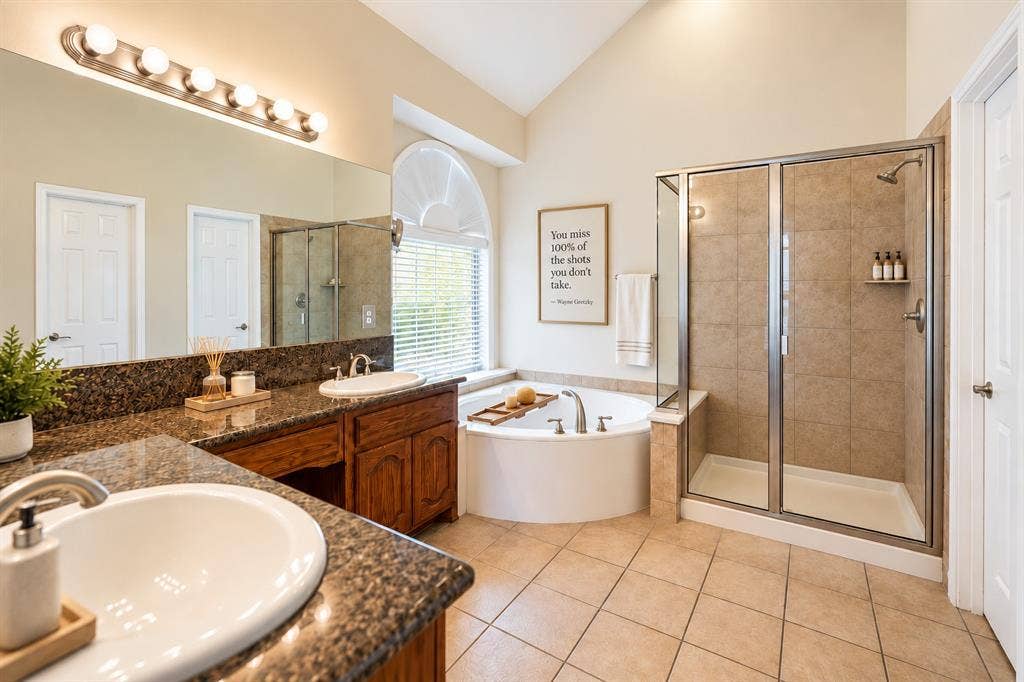 1736 Blue Stream Drive, DeSoto, TX 75115 Bathroom featuring a dual-sink vanity with stone countertops, a corner soaking tub, a glass-enclosed shower, tile flooring, and an arched window *Virtually staged to show potential*