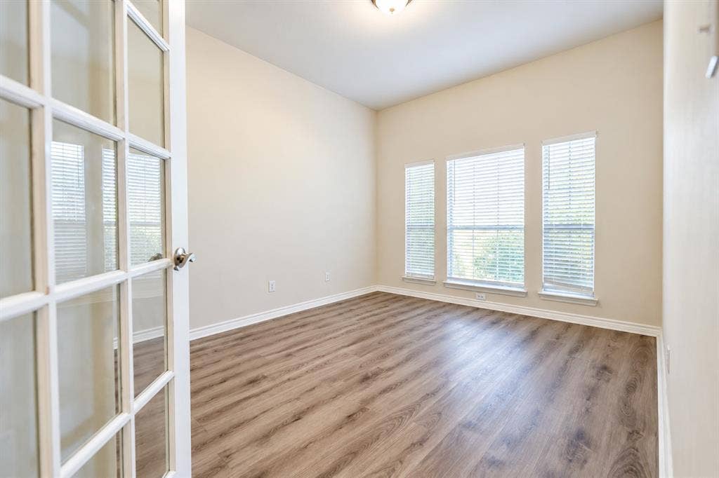 1736 Blue Stream Drive, DeSoto, TX 75115 Room featuring wood-look LVP flooring, neutral wall paint, three large windows with horizontal blinds, and a glass-paneled French door
