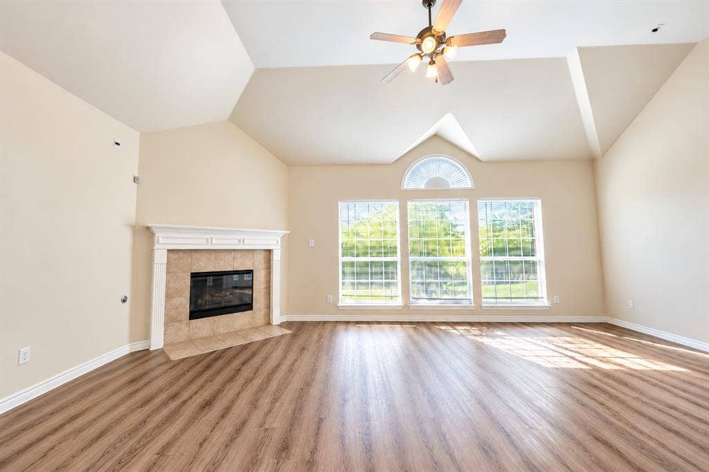 1736 Blue Stream Drive, DeSoto, TX 75115 Vaulted ceiling with an integrated multi-light ceiling fan, wood-look LVP flooring, and a fireplace with a white mantel and tile surround, accented by large windows featuring a semi-circular transom