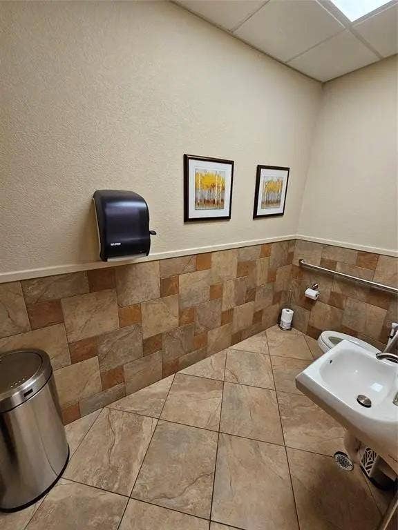 1831 W Louisiana Street, McKinney, TX 75069 Restroom featuring neutral wall-to-wall stone-finish tile, mounted grab bar, wall-mounted sink, and drop-ceiling tiles