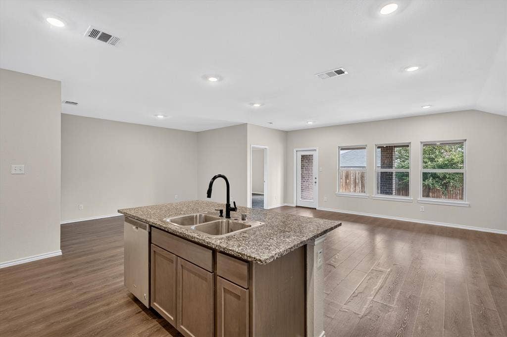 114 Real Quiet Lane, Caddo Mills, TX 75135 Kitchen island featuring a stone-look countertop, undermount stainless steel double-basin sink, matte black gooseneck faucet, and wood-finish cabinetry, complemented by wood-finish flooring and recessed lighting